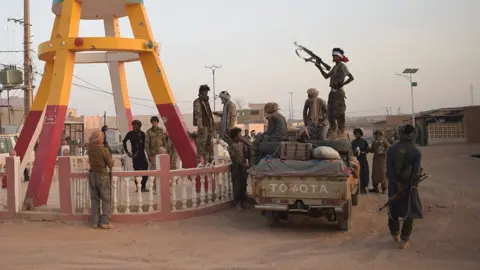 Des combattants en treillis, armés de fusils automatiques, debout autour d'un pick-up sur une route poussiéreuse. Un homme en silhouette se tient sur le véhicule, tenant un fusil. À gauche, les pattes d'une sculpture en bois aux couleurs brunes et jaunes sont visibles.